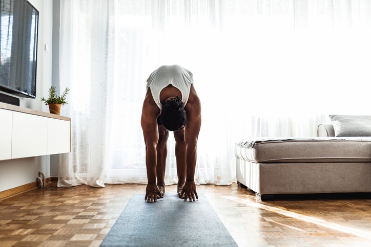 A man stretches his back and hamstrings by reaching for his toes while standing at his home during the day.